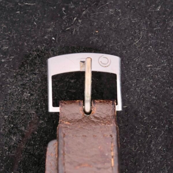 Close-up of a brown leather strap with a silver metal buckle, placed on a textured dark surface. Reminiscent of the Certina Club 2000 Cal. 2502 style, the buckle features a small circular logo on its upper edge.