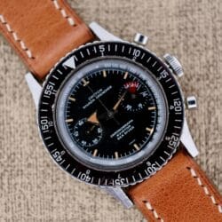 A vintage Nivada Grenchen Aviator Sea Diver Chronograph 1st Gen wristwatch with a black dial and brown leather strap rests elegantly on a textured surface.