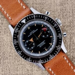 A vintage Nivada Grenchen Aviator Sea Diver Chronograph 1st Gen wristwatch with a black dial and brown leather strap rests elegantly on a textured surface.