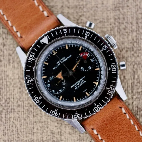 A vintage Nivada Grenchen Aviator Sea Diver Chronograph 1st Gen wristwatch with a black dial and brown leather strap rests elegantly on a textured surface.