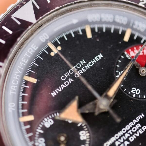 Close-up of a vintage Nivada Grenchen Aviator Sea Diver Chronograph 1st Gen watch face with a black dial, tachymeter on the outer rim, two subdials, luminous hands and markers, and a red-tipped second hand partially visible.