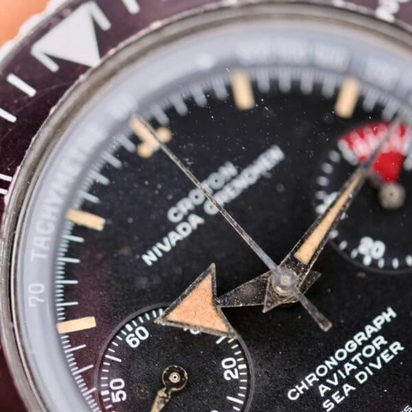A vintage Nivada Grenchen Aviator Sea Diver Chronograph 1st Gen watch close-up shows a dusty glass, a black dial with beige hands, a red subdial, and white markers. Text includes Tachymeter, Chronograph, Aviator, and Sea Diver.
