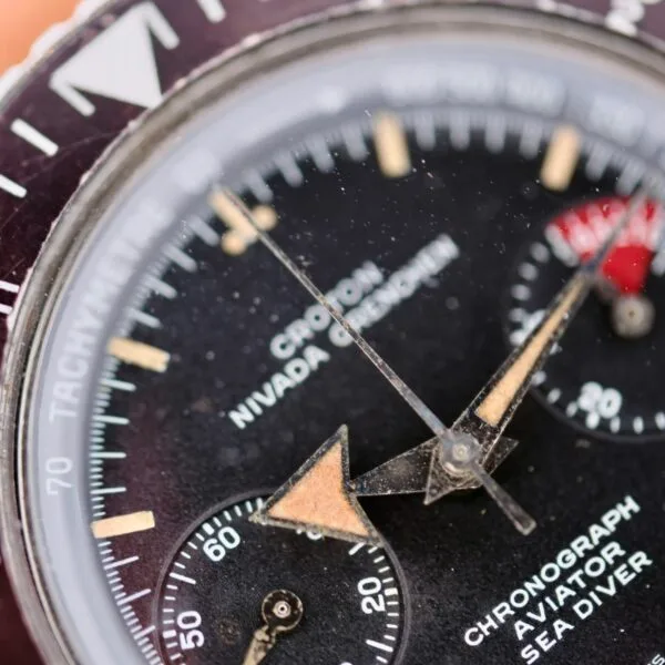 A vintage Nivada Grenchen Aviator Sea Diver Chronograph 1st Gen watch close-up shows a dusty glass, a black dial with beige hands, a red subdial, and white markers. Text includes Tachymeter, Chronograph, Aviator, and Sea Diver.
