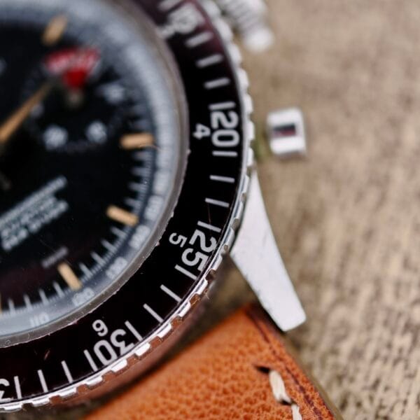 A close-up of the Nivada Grenchen Aviator Sea Diver Chronograph 1st Gen wristwatch reveals a black dial with a silver bezel and numbers, luminescent markers, and silver hands. It features a light brown leather strap with smooth stitching against a blurred textured background.
