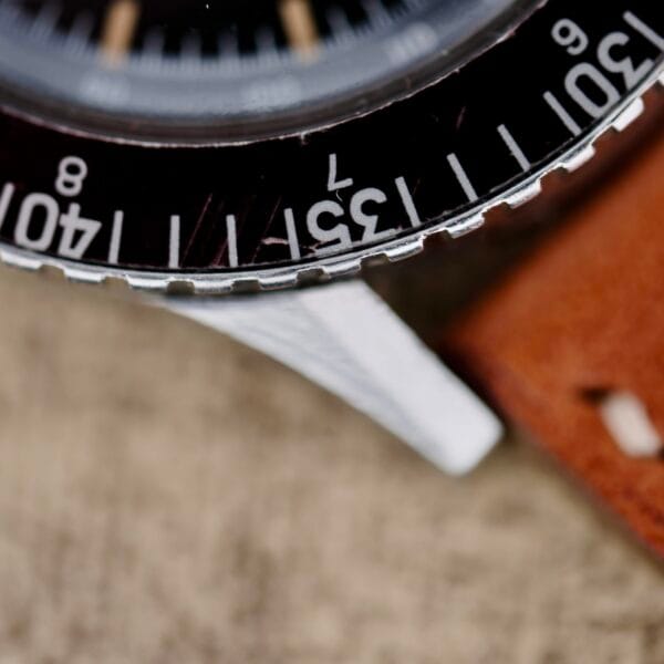A close-up of the Nivada Grenchen Aviator Sea Diver Chronograph 1st Gen showcases a black rotating bezel with numerals 30 and 40. The sleek silver case contrasts beautifully with a brown leather strap, set against a blurred background that highlights this elegant timepieces details.