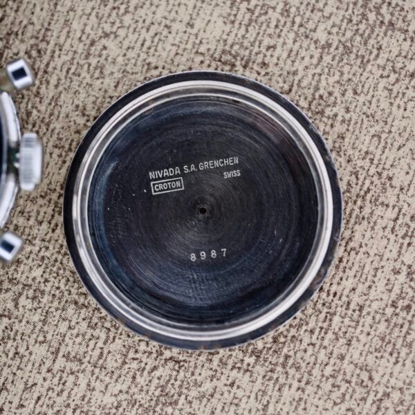 A close-up of the inside case back of a Nivada Grenchen Aviator Sea Diver Chronograph 1st Gen, showing engraved text and the number 8987, on a textured beige surface.