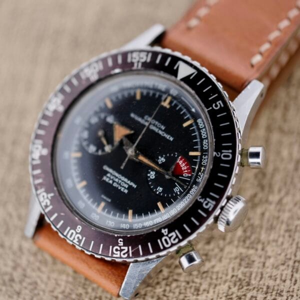Close-up of a vintage Nivada Grenchen Aviator Sea Diver Chronograph 1st Gen with a black dial, three subdials, and tachymeter bezel. It features silver-toned hands and markers on a textured beige surface and comes with a tan leather strap.