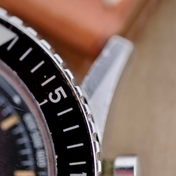 Close-up of the Nivada Grenchen Aviator Sea Diver Chronograph 1st Gen bezel showing numbers 5 and partial 6. The black bezel with white markings contrasts with the metallic case for a timeless look. The photo emphasizes the edge, artistically blurring background details.