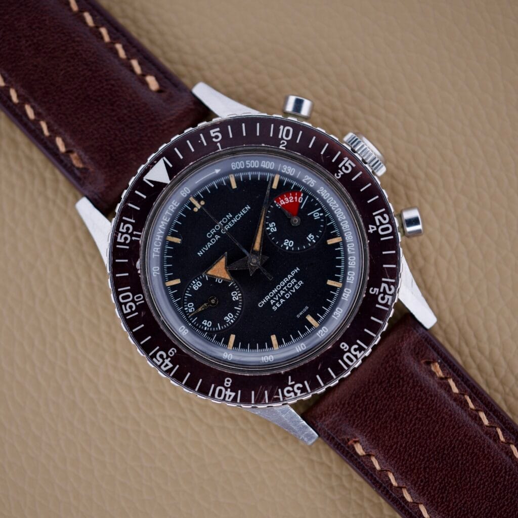 A Nivada Grenchen Aviator Sea Diver Chronograph 1st Gen with a brown leather strap rests elegantly on a tan textured surface.