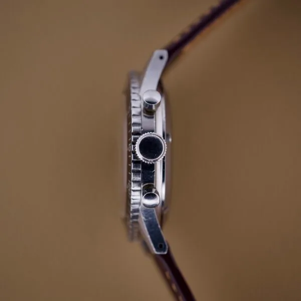 Side view of a silver Nivada Grenchen Aviator Sea Diver Chronograph 1st Gen wristwatch with a brown leather strap, set against a plain tan background.