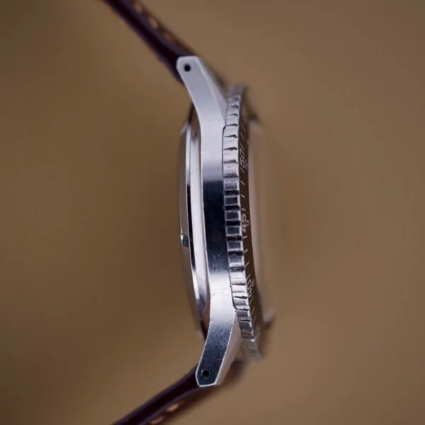 Side view of a Nivada Grenchen Aviator Sea Diver Chronograph 1st Gen wristwatch with a metal case, grooved bezel, and brown leather strap against a beige background.