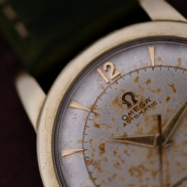 Close-up of a vintage Omega Seamaster Gold Automatic 354 wristwatch with a weathered face, marked dial, and patina. Features gold hour markers and hands, the number 12 in gold at the top, and the Omega logo beneath it.