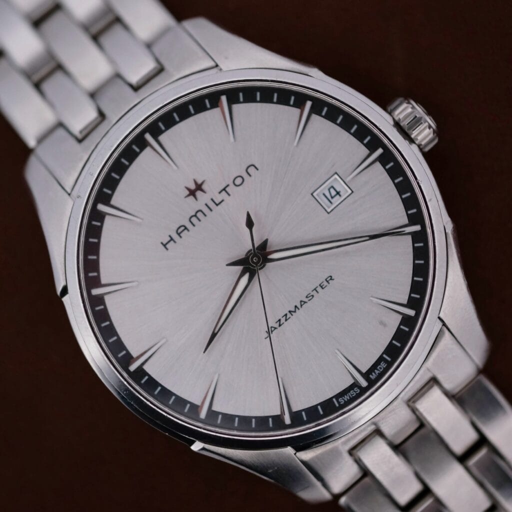 On a dark background, the Hamilton Jazz Master Stainless Steel wristwatch gleams with its silver face, black hour markers, sleek black hands, and a date window displaying 14, complemented by a stainless steel bracelet.