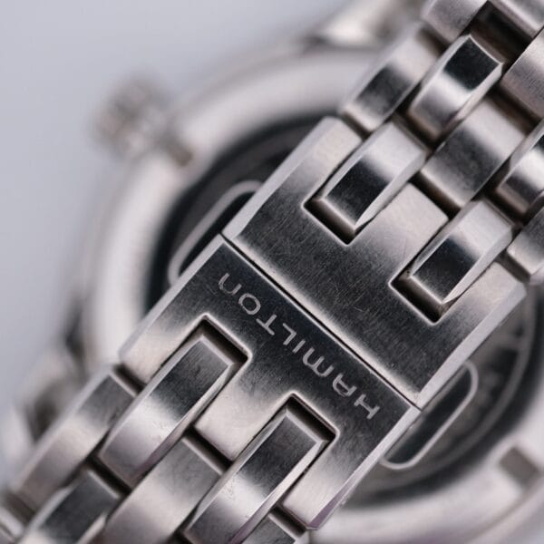 Close-up of the Hamilton Jazz Master Stainless Steel watch bracelet with interlocking links, showcasing timeless elegance. The clasp features an engraved Hamilton, and the softly blurred background highlights the bracelets exquisite texture and design.