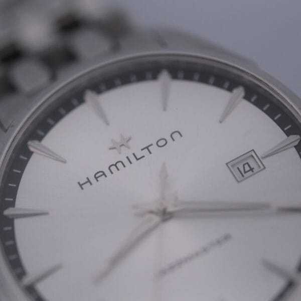 Close-up of a Hamilton Jazz Master Stainless Steel watch highlighting the silver dial with shiny hour markers, a date window showing 14, and a partly visible minute hand. The Hamilton logo is prominently shown below the 12 oclock position.