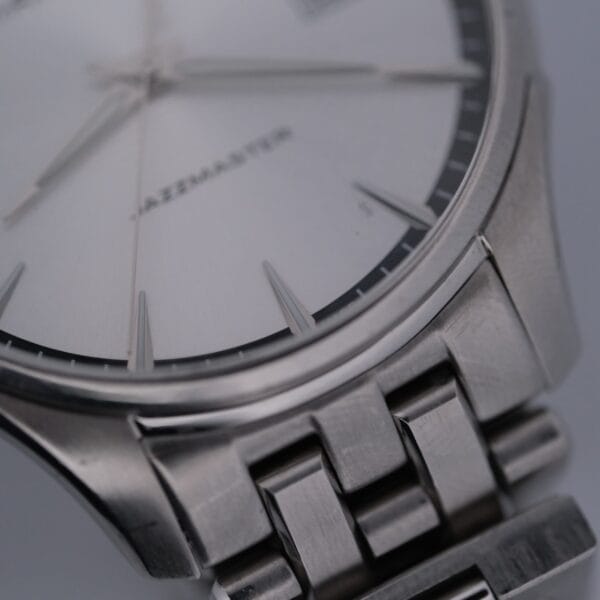 Close-up of the Hamilton Jazz Master Stainless Steel watch with a metallic bracelet, showcasing a minimalist design with slender hour markers on a white dial. The word JAZZMASTER is partially visible on the watch face.