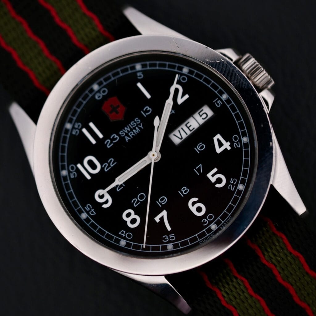 Close-up of the Swiss Army Day/Date Field Watch Swiss Made, showcasing a black face with white numerals and hands. It displays Vie, 5 for the day and date, features a black and red striped fabric strap, and is accented with a silver-toned bezel.