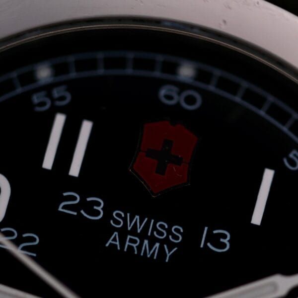 Close-up of a Swiss Army Day/Date Field Watch Swiss Made, showcasing a black dial with white hour markers and numbers. The iconic red and white cross emblem is at the center. The numbers 23 and 13 are visible, along with the text SWISS ARMY.
