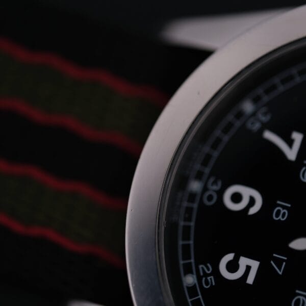 Close-up of the Swiss Army Day/Date Field Watch, Swiss Made, showcasing a black dial with large white numerals and a metal casing. It features minute and second marks for precision and comes with a black and red-striped fabric strap.