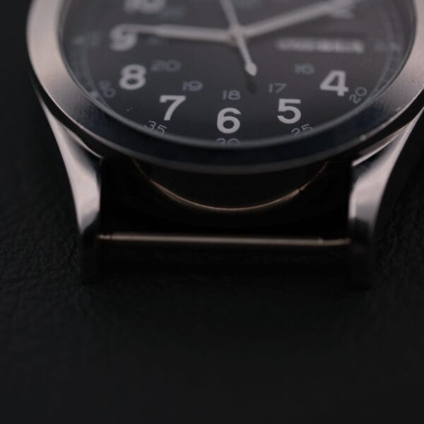 Close-up of the Swiss Army Day/Date Field Watch Swiss Made, featuring a black dial with white numerals and a partially visible day/date window. The metallic case and strap attachment subtly stand out against a dark backdrop.