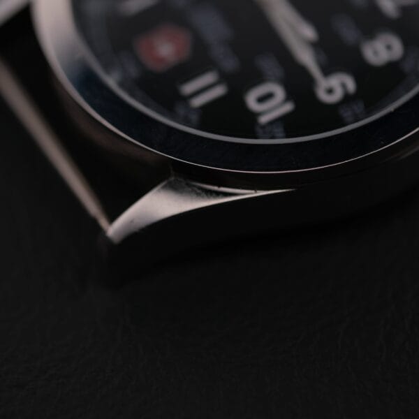 Close-up of the Swiss Army Day/Date Field Watch Swiss Made, featuring a black dial with white numbers and a shiny metal frame. The image highlights the watch case edge against a dark background, emphasizing its sleek design.