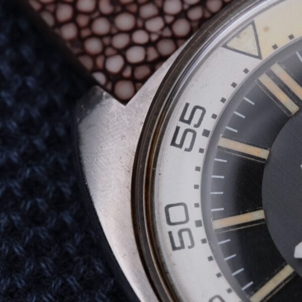 Close-up of a Bulova Accutron Deep Sea 666 “Devil Diver” Inner Bezel wristwatch featuring a metal case and brown textured strap. The detailed watch face includes markings like numbers 50 and 55 on the outer edge, set against a dark blue textured fabric backdrop, enhancing its elegance.
