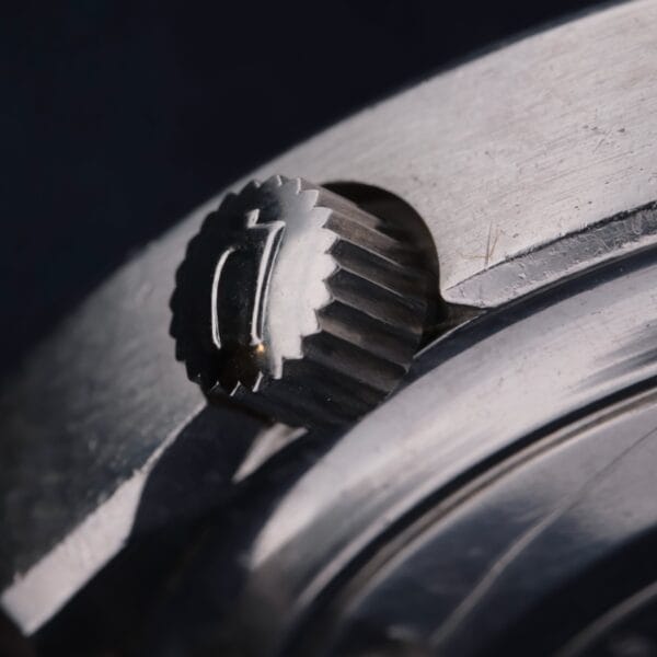 Close-up of a metallic watch crown with grooved edges and an engraved symbol on top, set against a blurred dark background. The crown belongs to the Bulova Accutron Deep Sea 666 “Devil Diver” Inner Bezel, showcasing intricate details and a brushed finish as part of its larger casing.