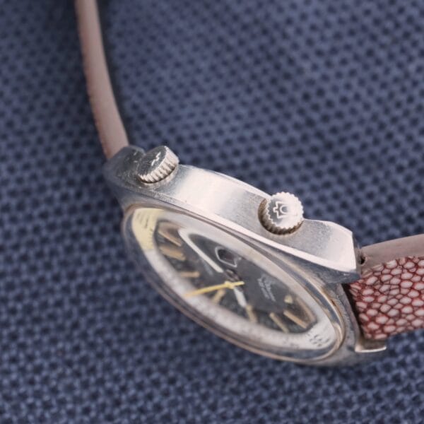A close-up of the vintage Bulova Accutron Deep Sea 666 Devil Diver Inner Bezel wristwatch with a dark face and metallic case rests on textured blue fabric, featuring two small crowns on its side and a brown leather strap with a reptile pattern.