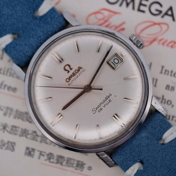 A close-up of the Omega Seamaster Deville Automatic for the Japanese Market showcases a silver dial with baton markers and a date display at 3 oclock. The watch features a blue leather strap, elegantly set against a background with text.