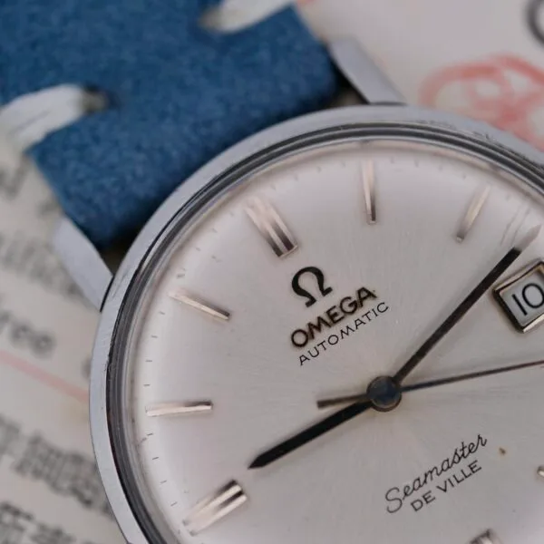 A close-up of the Omega Seamaster Deville Automatic for the Japanese Market highlights its elegant silver face and blue leather strap. The date shows the 10th, featuring gleaming silver hour and minute hands. The distinctive Omega branding is beautifully displayed on the dial.