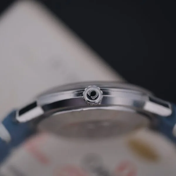 Close-up of the Omega Seamaster Deville Automatic crown, exclusive to the Japanese market, with intricate detailing against a blurred backdrop. It boasts a blue strap and sleek metal case, exemplifying craftsmanship and style.