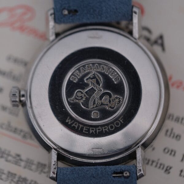 Close-up of an Omega Seamaster Deville Automatic for the Japanese market, showcasing an embossed seahorse logo and Seamaster Waterproof text on the back. The watch, featuring a blue strap, is displayed on a text-covered surface.