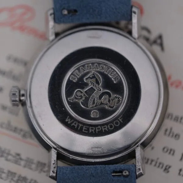 Close-up of an Omega Seamaster Deville Automatic for the Japanese market, showcasing an embossed seahorse logo and Seamaster Waterproof text on the back. The watch, featuring a blue strap, is displayed on a text-covered surface.