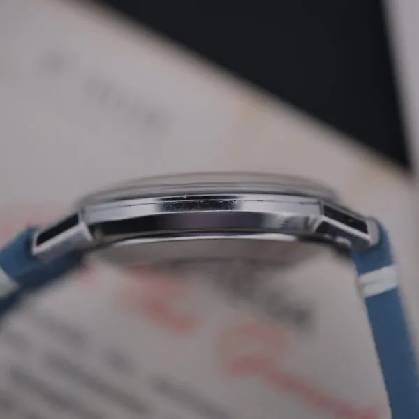 Close-up of an Omega Seamaster Deville Automatic for the Japanese Market, featuring a blue strap and round silver-colored case; blurred background with indistinct text on paper.