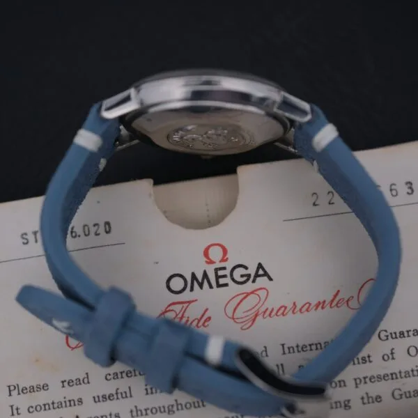 A silver Omega Seamaster Deville Automatic, designed for the Japanese market, features a blue leather strap and elegantly rests on a certificate marked Guarantee, partially visible. The dark backdrop beautifully emphasizes both the watch and document.