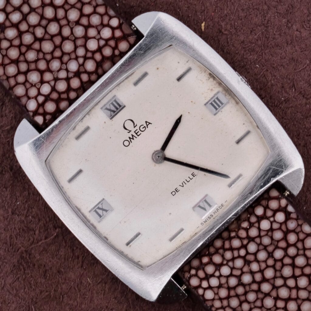 The Omega Deville TV Case Stainless Steel watch boasts a silver square face with black hands and Roman numerals at 12, 3, 6, and 9 oclock. It features a textured brown leather strap against a dark brown background, reminiscent of iconic TV case designs.