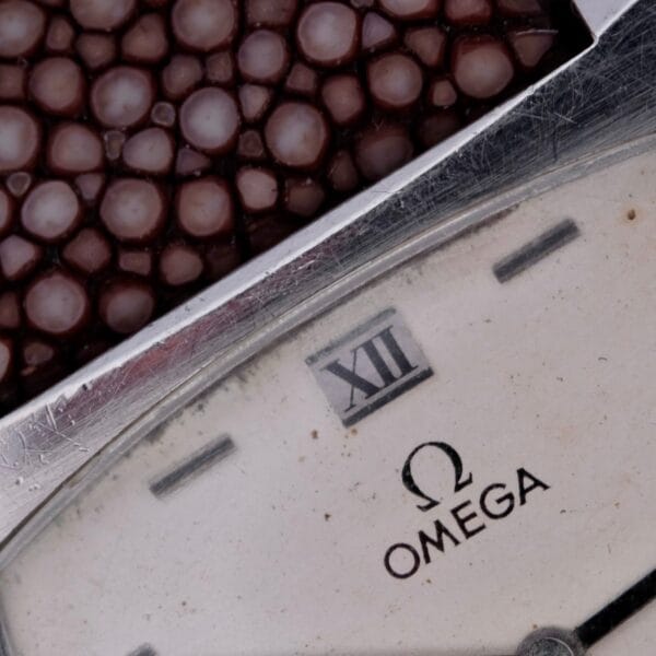 Close-up of an Omega Deville TV Case Stainless Steel wristwatch with a distinctive TV case and Roman numeral XII on the dial. A textured brown strap is shown at the top, while slight scuffs on the watch face narrate its history of wear.