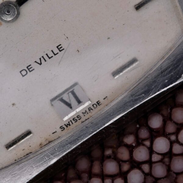 Close-up of the Omega Deville TV Case Stainless Steel watch face, with the distinctive Swiss Made mark. The Roman numeral VI decorates the six oclock position within its refined TV case. Resting on a textured reddish-brown surface, this vintage timepiece exudes timeless charm.