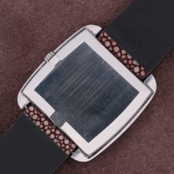 A close-up of an Omega Deville TV Case Stainless Steel watch with a scratched silver case and square, reflective face, resting on a brown textured background, featuring a black leather strap with brown detailing near the case.