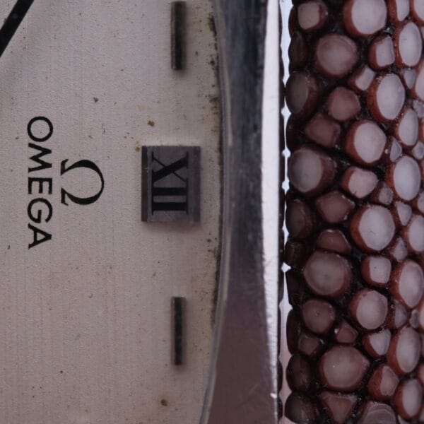 Close-up of an Omega Deville TV Case Stainless Steel watch face, showcasing the Roman numeral XII and Omega logo. The side features a distinctive textured design with brown and white round stones or beads, adding unique decoration.