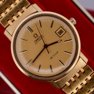 A close-up of the Omega Deville Automatic Gold Tone Date Full Set wristwatch highlights its matching bracelet, black hour markers, a date window showing 26 at 3 oclock, and the iconic Omega logo with AUTOMATIC and DE VILLE.