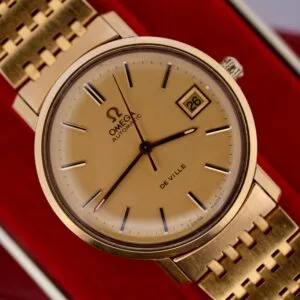 A close-up of the Omega Deville Automatic Gold Tone Date Full Set wristwatch highlights its matching bracelet, black hour markers, a date window showing 26 at 3 oclock, and the iconic Omega logo with AUTOMATIC and DE VILLE.
