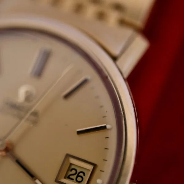 Close-up of the Omega Deville Automatic Gold Tone Date Full Set wristwatch, featuring part of the dial with silver hour markers and hands. The date window shows 26th against a soft red background, enhancing its timeless elegance.