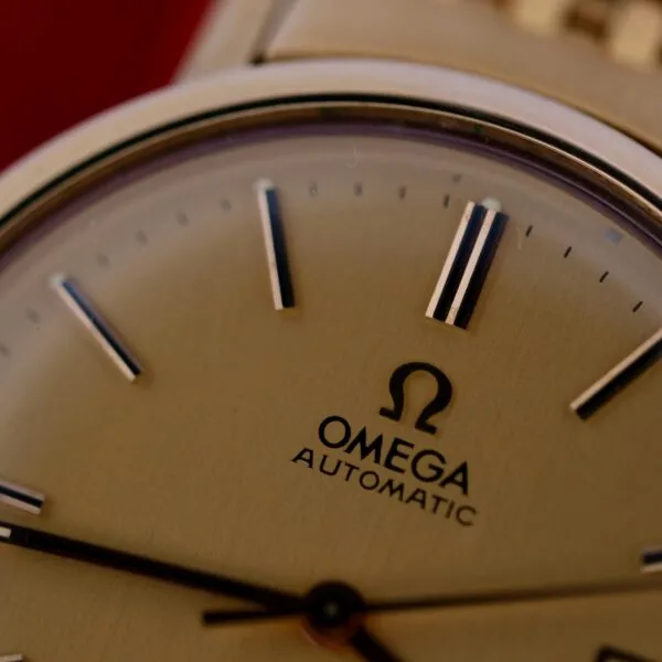 Close-up of the Omega Deville Automatic Gold Tone Date Full Set watch face, featuring a classic design with a beige background, simple hour markers, and the Omega logo above AUTOMATIC. Metallic hands complement the elegant watch face.