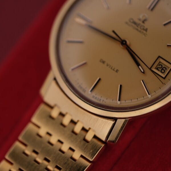 Close-up of an Omega Deville Automatic Gold Tone Date Full Set wristwatch, featuring a round face with date 26, gold bracelet, thin hour markers, and brand name at the top. The elegant design is displayed against a striking red background.