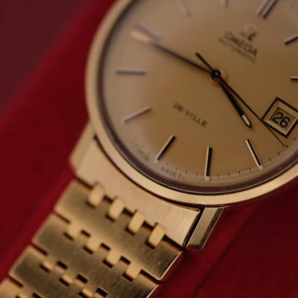 Close-up of an Omega Deville Automatic Gold Tone Date Full Set wristwatch, featuring a round face with date 26, gold bracelet, thin hour markers, and brand name at the top. The elegant design is displayed against a striking red background.