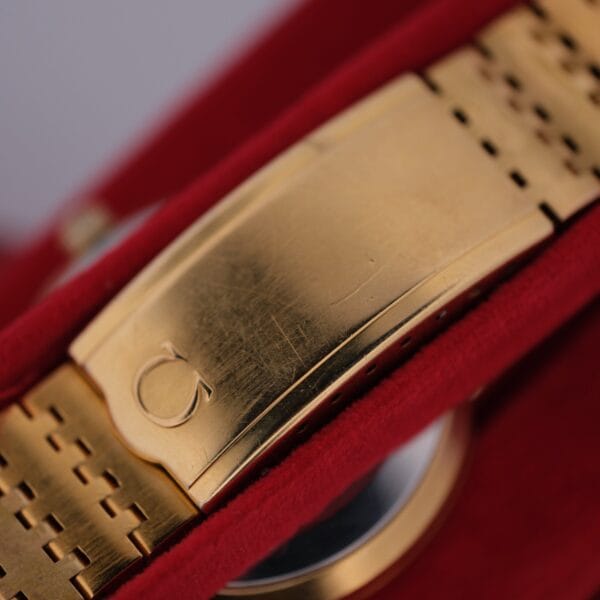 An Omega Deville Automatic Gold Tone Date Full Set watch is showcased on a red velvet surface, emphasizing its brushed metallic texture and intricate bracelet links, with a distinctive clasp engraved with the logo.