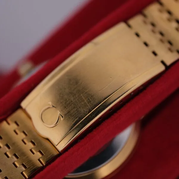 An Omega Deville Automatic Gold Tone Date Full Set watch is showcased on a red velvet surface, emphasizing its brushed metallic texture and intricate bracelet links, with a distinctive clasp engraved with the logo.