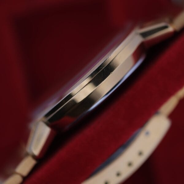 Close-up view of an Omega Deville Automatic Gold Tone Date Full Set wristwatch on a red background, showcasing the polished metal watch case edge and part of the beige strap.