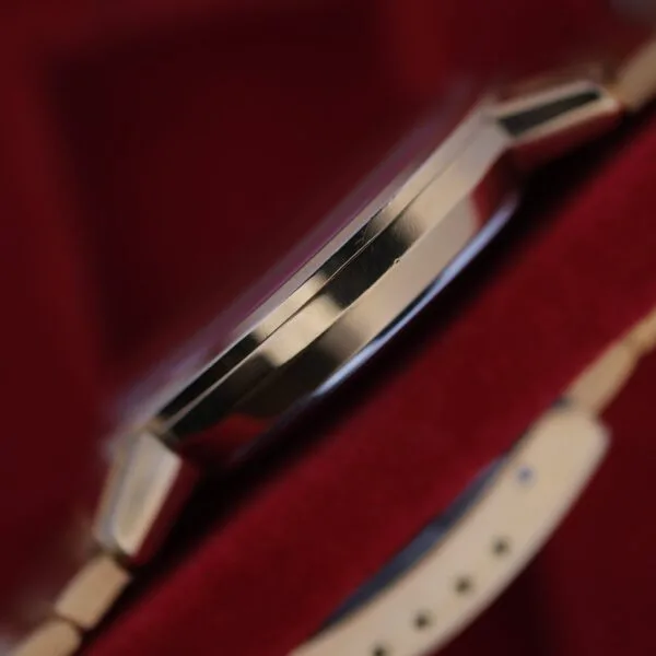 Close-up view of an Omega Deville Automatic Gold Tone Date Full Set wristwatch on a red background, showcasing the polished metal watch case edge and part of the beige strap.